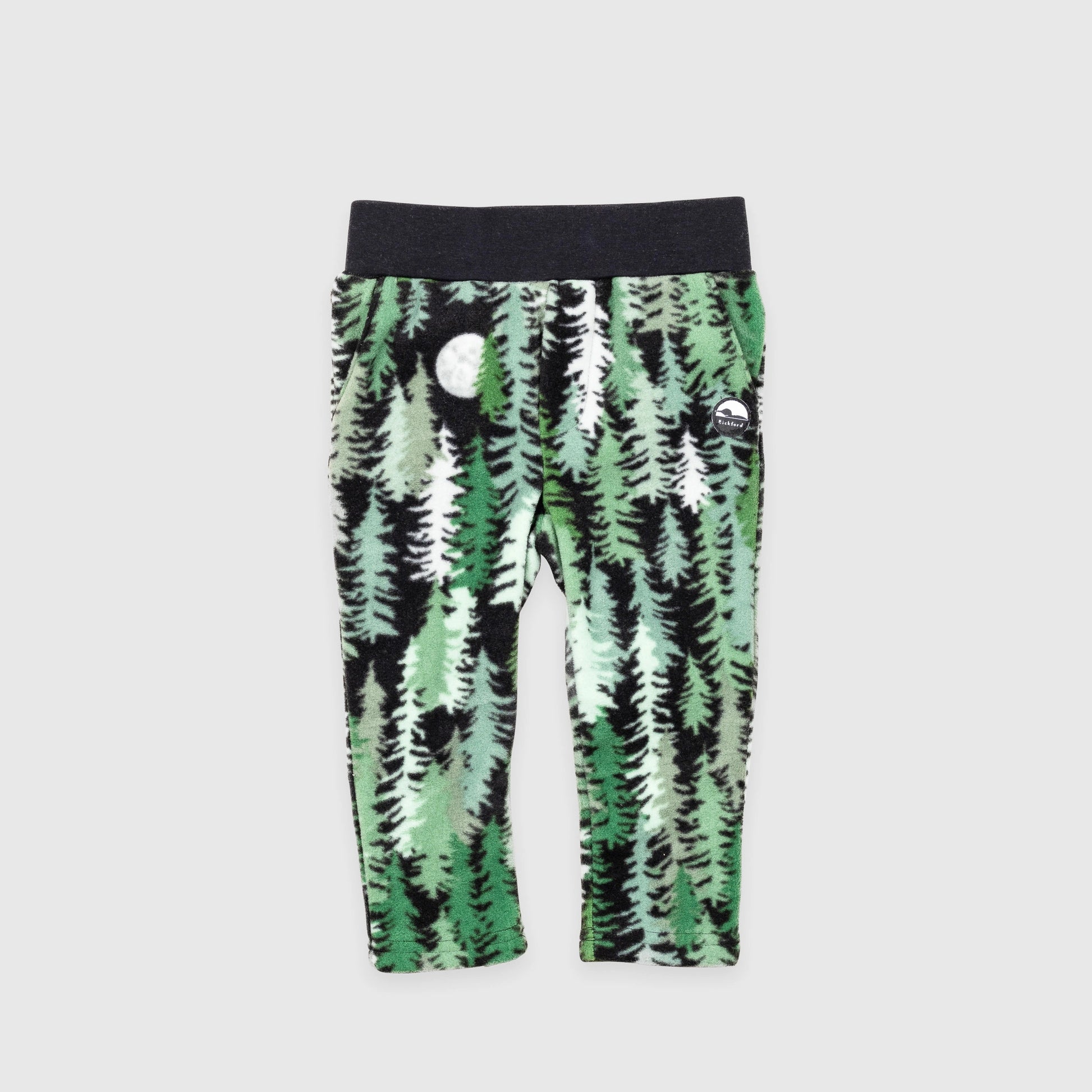 Bickford USA Youth fleece pants in the Eastern Pine colorway, laid flat on a light gray background, featuring an all-over print of layered pine trees in shades of green, black, and cream with a moon motif, with a wide black elastic waistband, side hand pockets, and circular Bickford logo patch on the hip.
