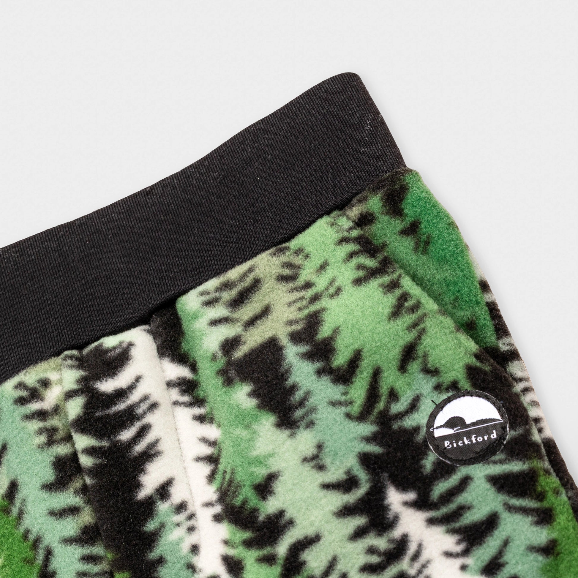 Close-up detail of the Eastern Pine Youth Pants showing the wide black elastic waistband, angled side pocket, and circular woven Bickford mountain logo patch against the green and black pine tree print.