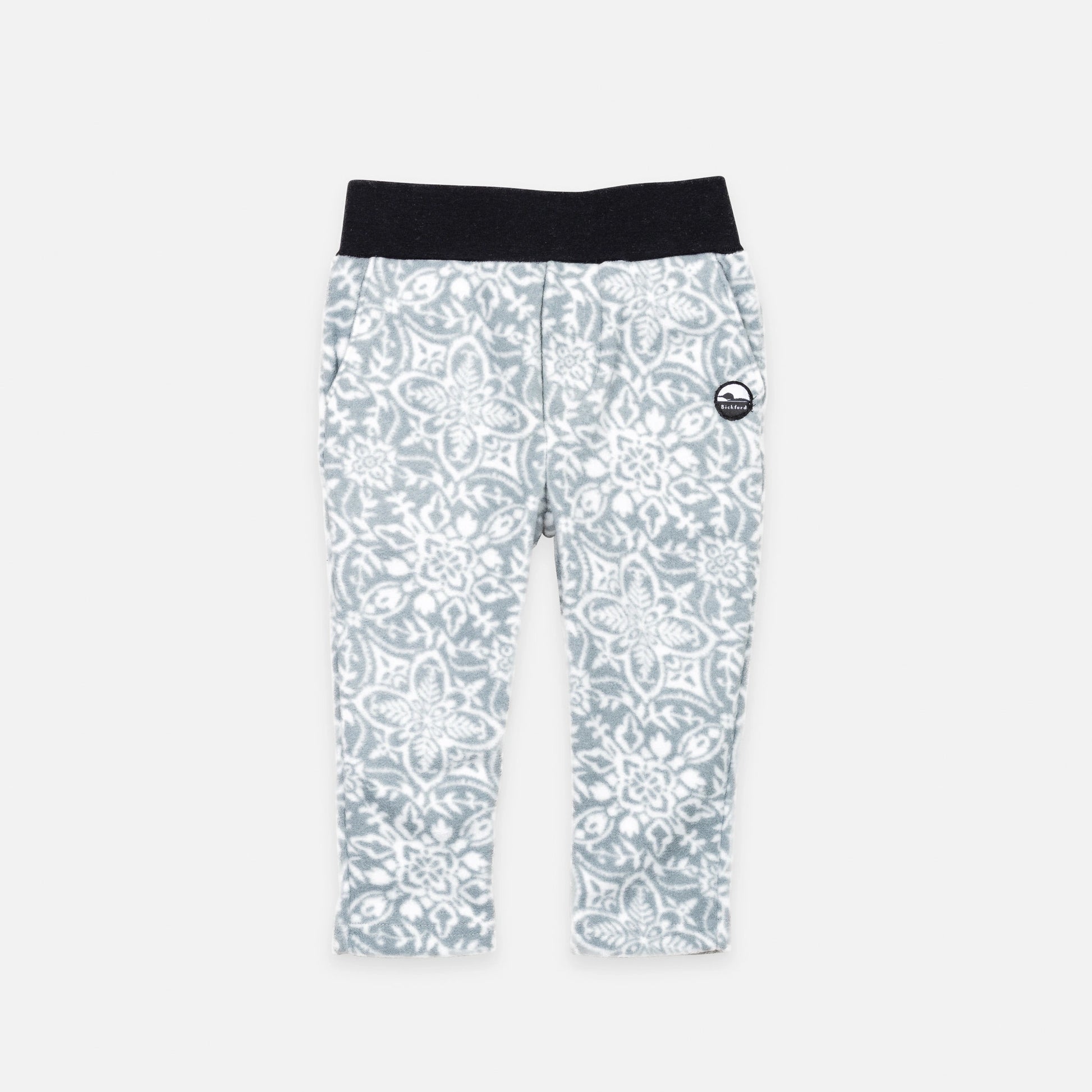 Bickford USA Youth fleece pants in the Snowflake colorway, laid flat on a light gray background, featuring an all-over ornate snowflake and medallion print in grey and white, with a wide black elastic waistband, side hand pockets, and circular Bickford logo patch on the hip.