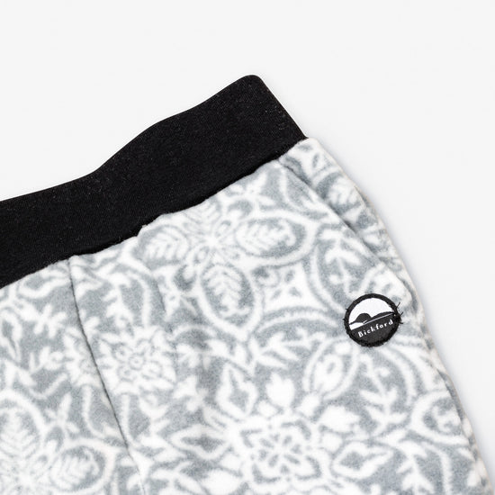 Close-up detail of the Snowflake Youth Pants showing the wide black elastic waistband, angled side pocket, and circular woven Bickford mountain logo patch against the grey and white ornate snowflake medallion pattern.