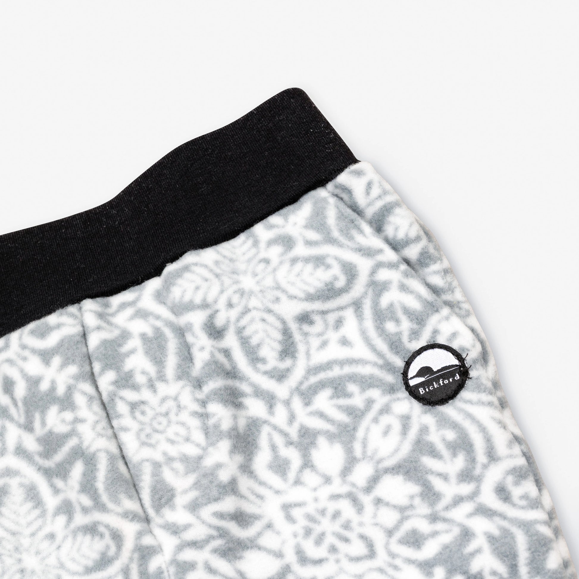 Close-up detail of the Snowflake Youth Pants showing the wide black elastic waistband, angled side pocket, and circular woven Bickford mountain logo patch against the grey and white ornate snowflake medallion pattern.