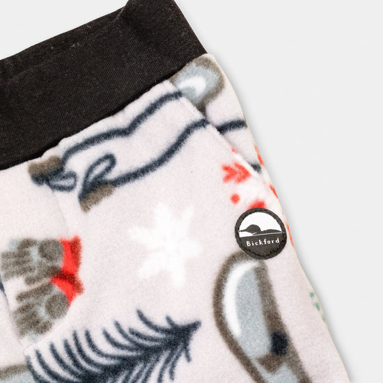 Close-up detail of the Sugarbush Youth Pants showing the wide black elastic waistband, angled side pocket, and circular woven Bickford mountain logo patch against the winter sports-themed print with skis, poles, snowflakes, and pine trees.