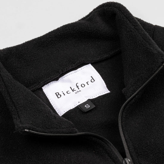 Close-up detail of the black Youth Quarter-Zip showing the open funnel collar with the Bickford USA label and size tag visible, and the metal zipper pull in black.