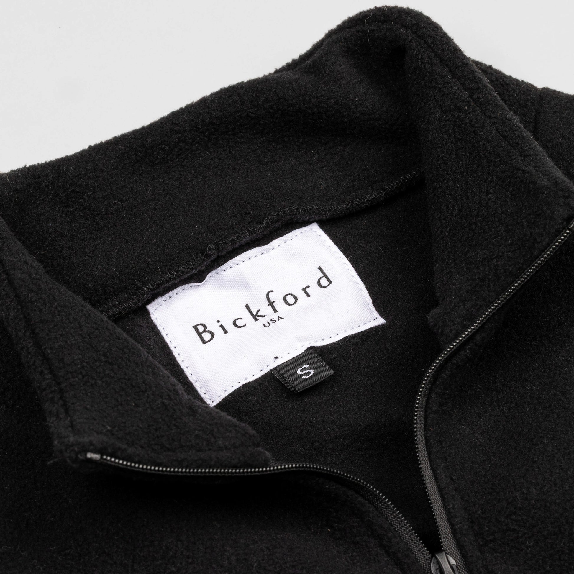 Close-up detail of the black Youth Quarter-Zip showing the open funnel collar with the Bickford USA label and size tag visible, and the metal zipper pull in black.
