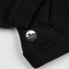 Close-up of the lower hem corner of the black Youth Quarter-Zip showing the circular woven Bickford logo patch with a mountain and horizon graphic.