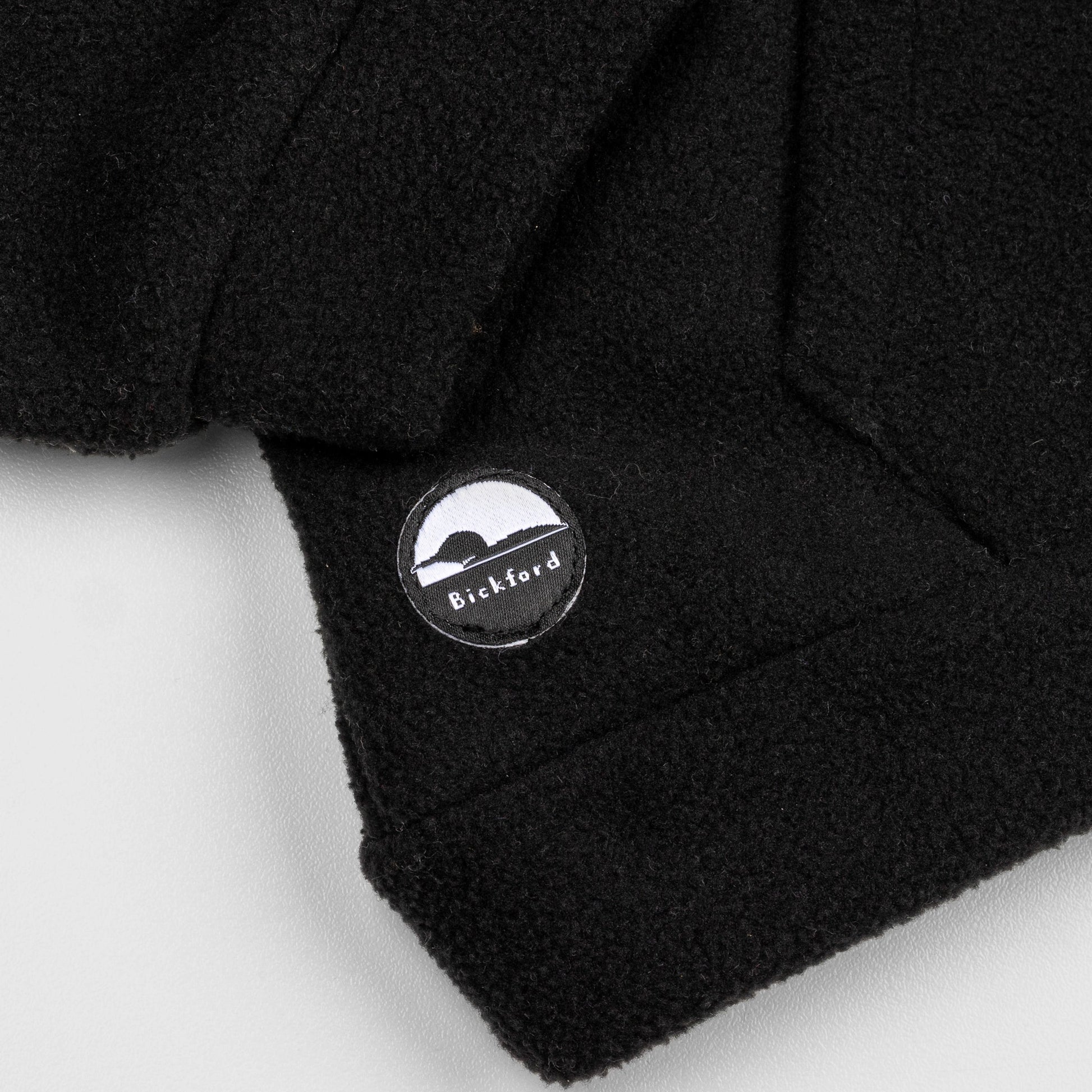 Close-up of the lower hem corner of the black Youth Quarter-Zip showing the circular woven Bickford logo patch with a mountain and horizon graphic.