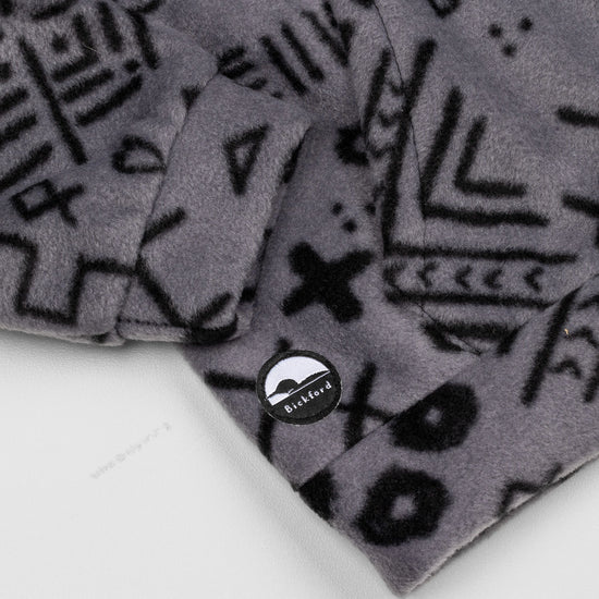 Close-up of the lower hem corner of the Boulder Youth Quarter-Zip showing the circular woven Bickford logo patch with a mountain graphic, set against the grey and black geometric print.