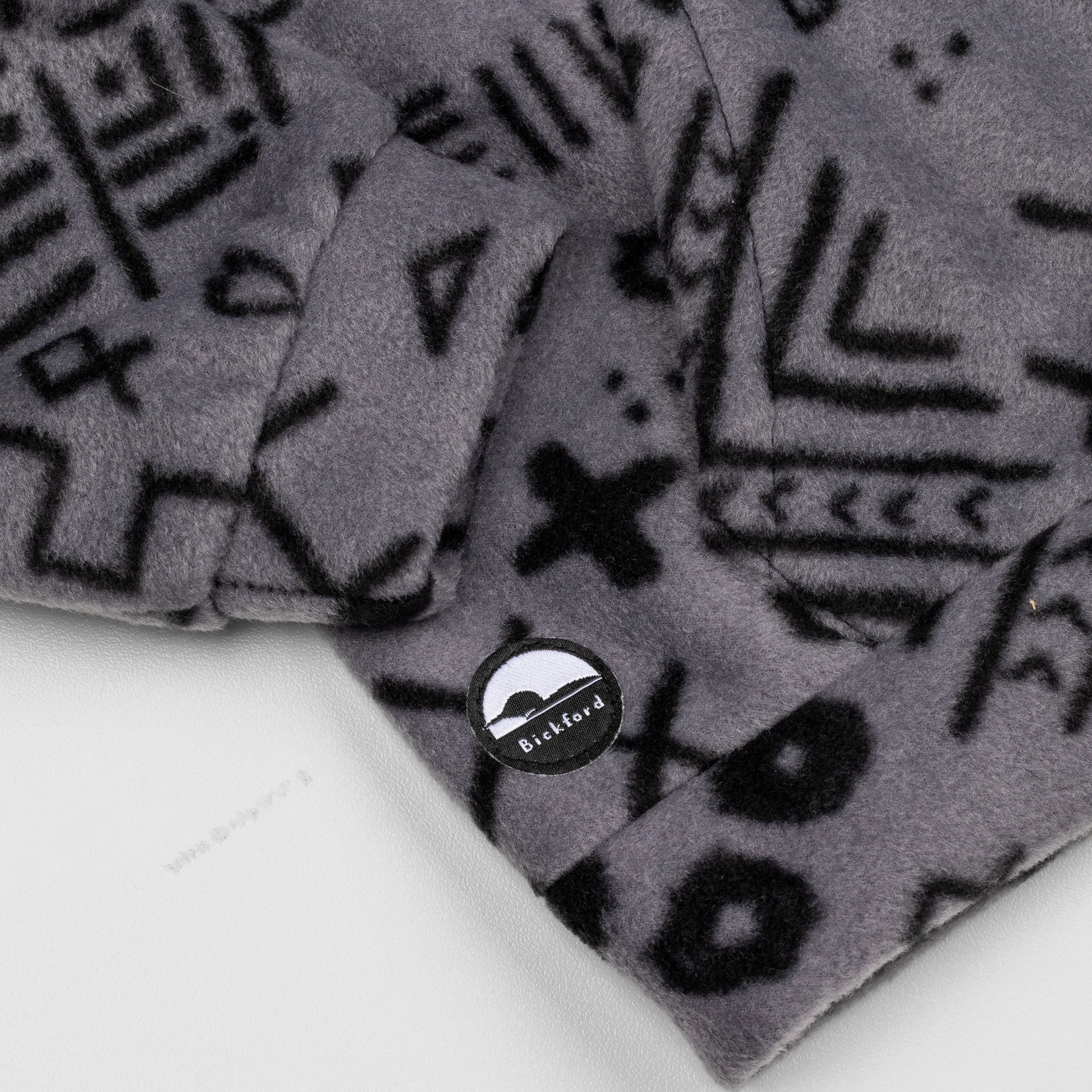 Close-up of the lower hem corner of the Boulder Youth Quarter-Zip showing the circular woven Bickford logo patch with a mountain graphic, set against the grey and black geometric print.