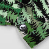 Close-up of the lower hem corner of the Eastern Pine Youth Quarter-Zip showing the circular woven Bickford logo patch with a mountain graphic, set against the green and black pine tree print.