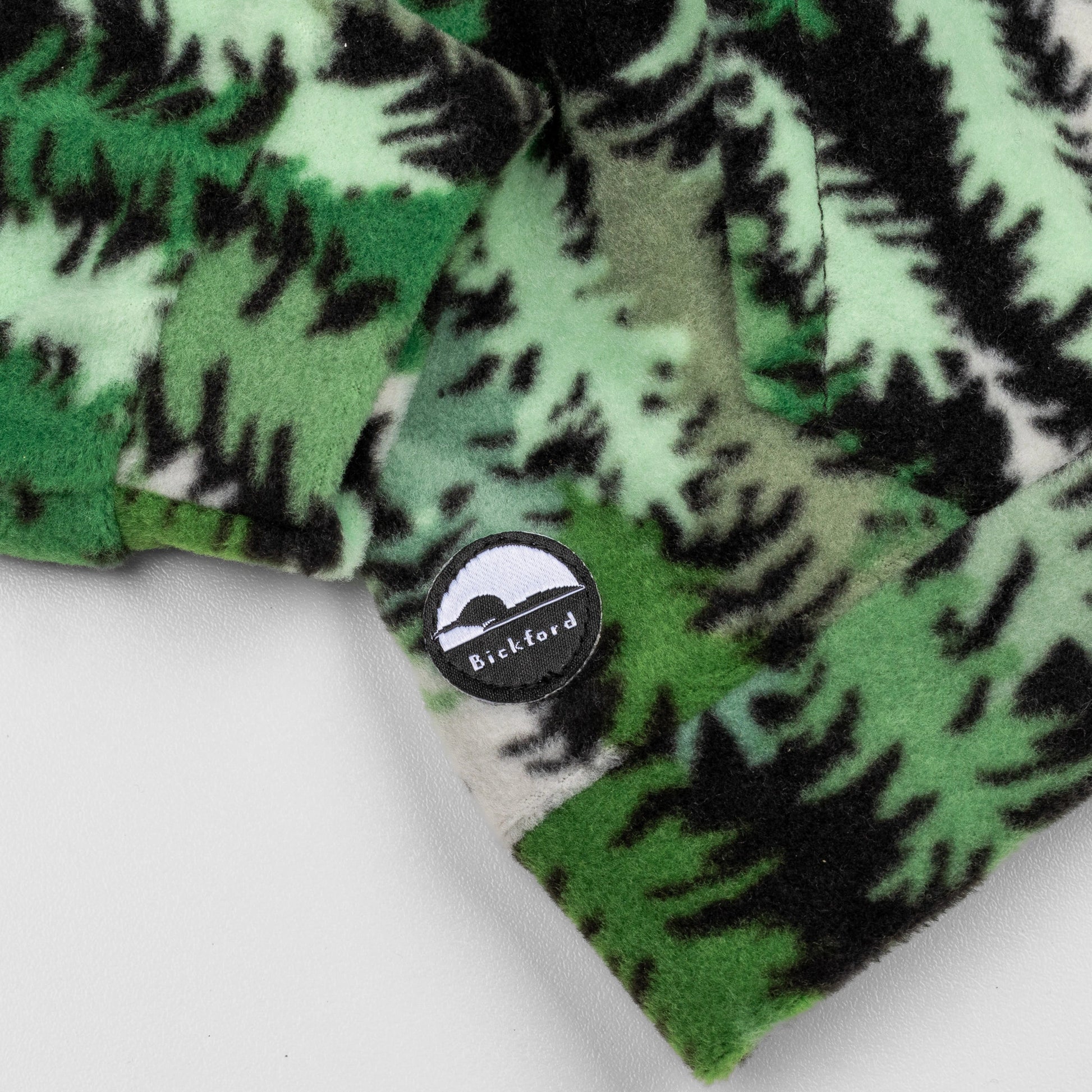 Close-up of the lower hem corner of the Eastern Pine Youth Quarter-Zip showing the circular woven Bickford logo patch with a mountain graphic, set against the green and black pine tree print.