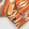 Close-up of the lower hem corner of the Heritage Trail Youth Quarter-Zip showing the circular woven Bickford logo patch with a mountain graphic, set against the southwestern geometric print in brown, orange, and grey.