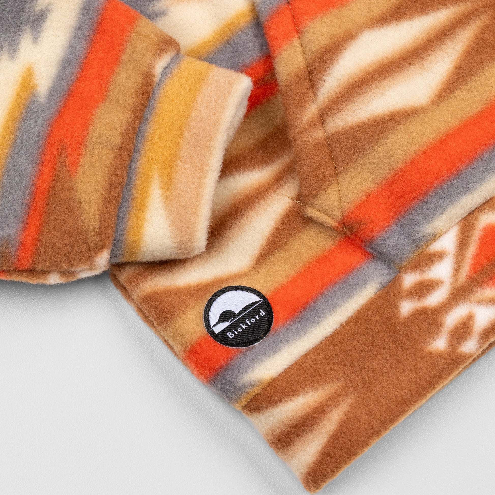 Close-up of the lower hem corner of the Heritage Trail Youth Quarter-Zip showing the circular woven Bickford logo patch with a mountain graphic, set against the southwestern geometric print in brown, orange, and grey.