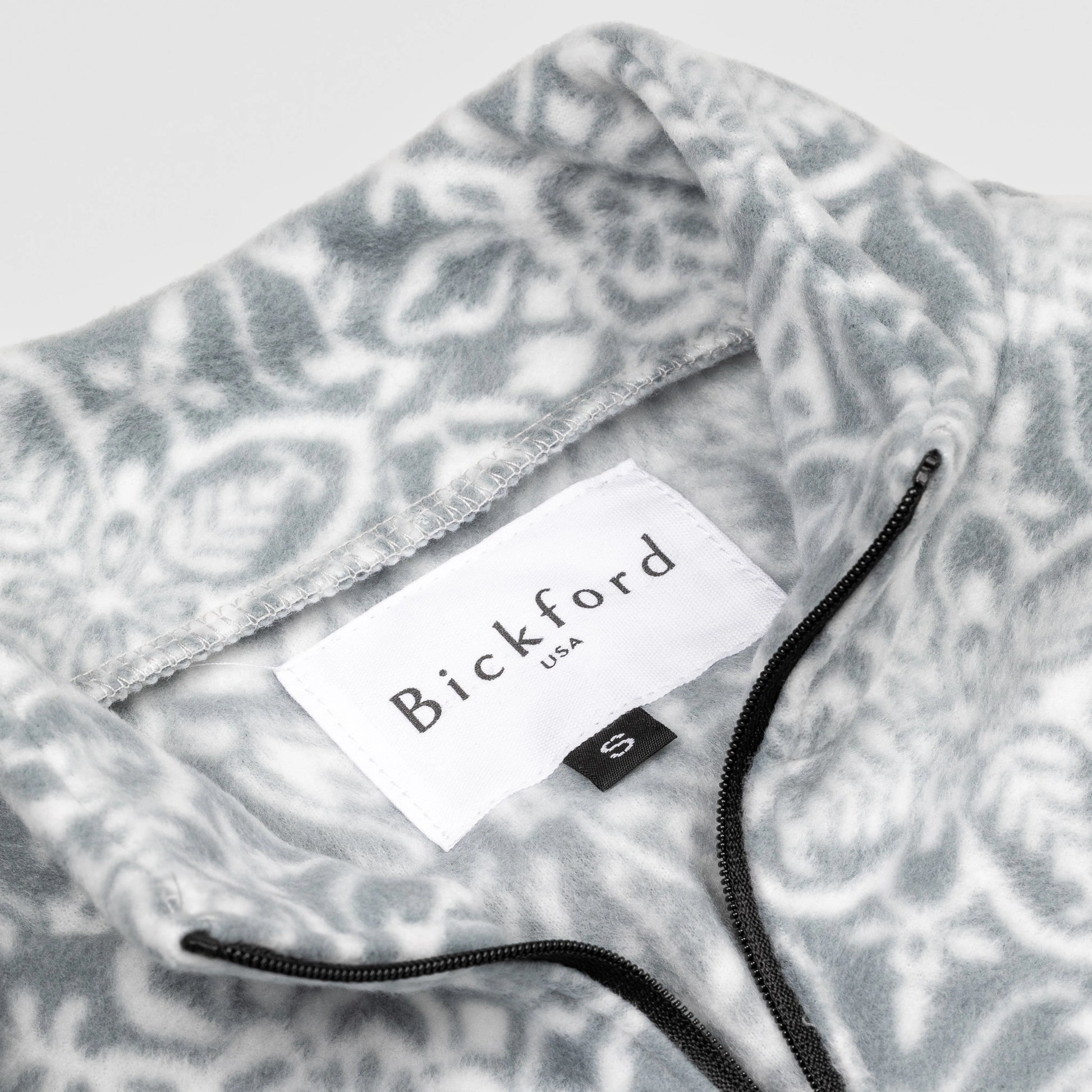 Close-up detail of the Snowflake Youth Quarter-Zip showing the open funnel collar with the Bickford USA label and size tag visible, with the grey and white medallion print visible on the inner collar and the black zipper.