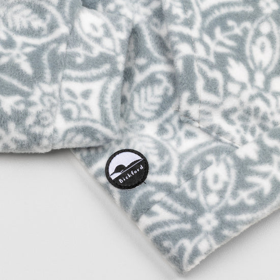 Close-up of the lower hem corner of the Snowflake Youth Quarter-Zip showing the circular woven Bickford logo patch with a mountain graphic, set against the grey and white ornate snowflake pattern.