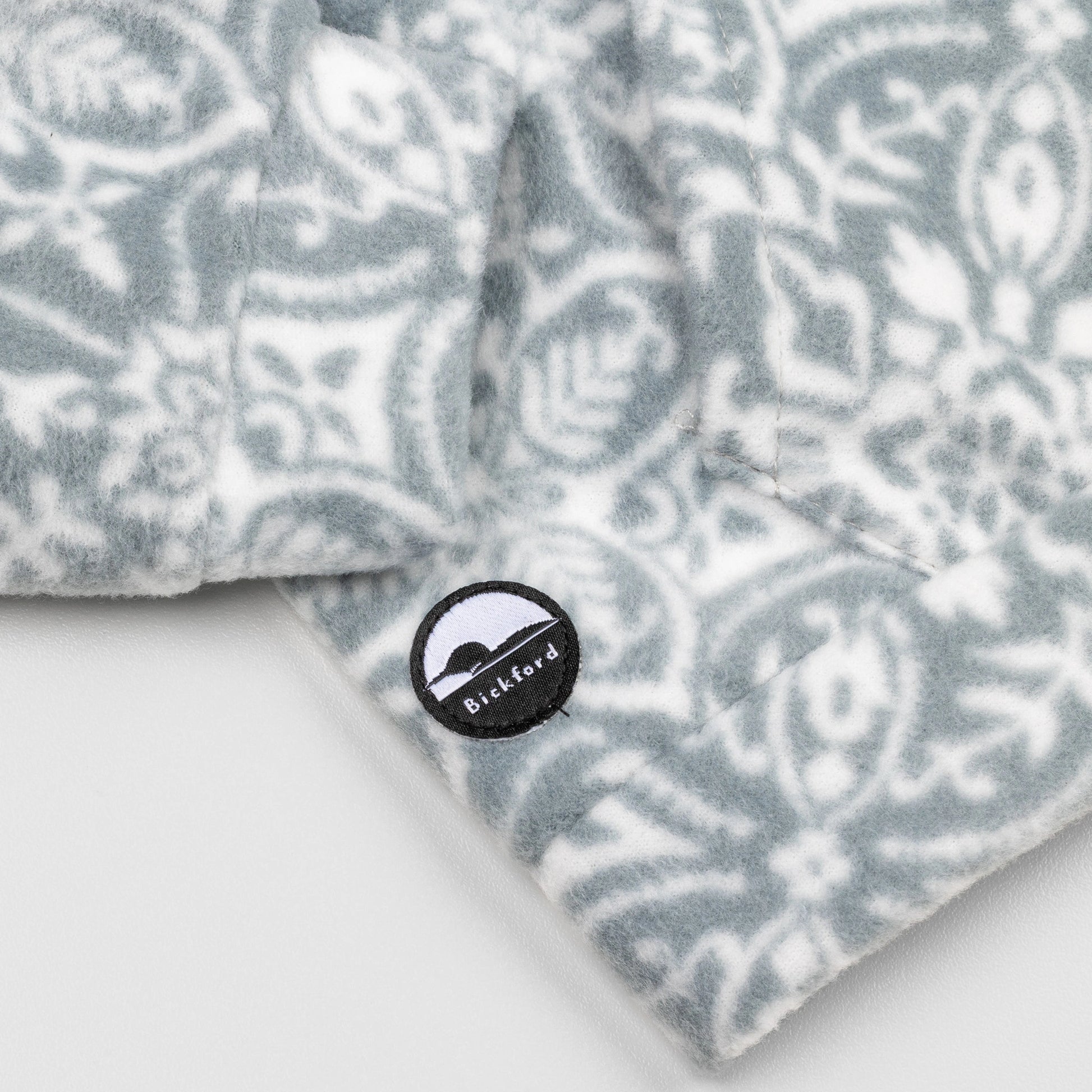 Close-up of the lower hem corner of the Snowflake Youth Quarter-Zip showing the circular woven Bickford logo patch with a mountain graphic, set against the grey and white ornate snowflake pattern.