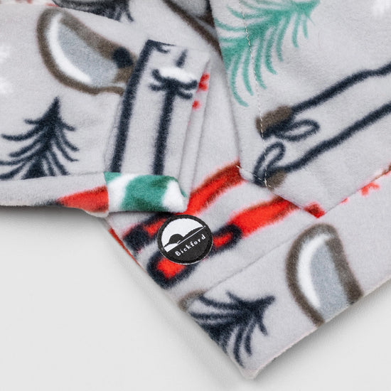 Close-up of the lower hem corner of the Sugarbush Youth Quarter-Zip showing the circular woven Bickford logo patch with a mountain graphic, set against the winter sports-themed print with skis, poles, and pine trees.