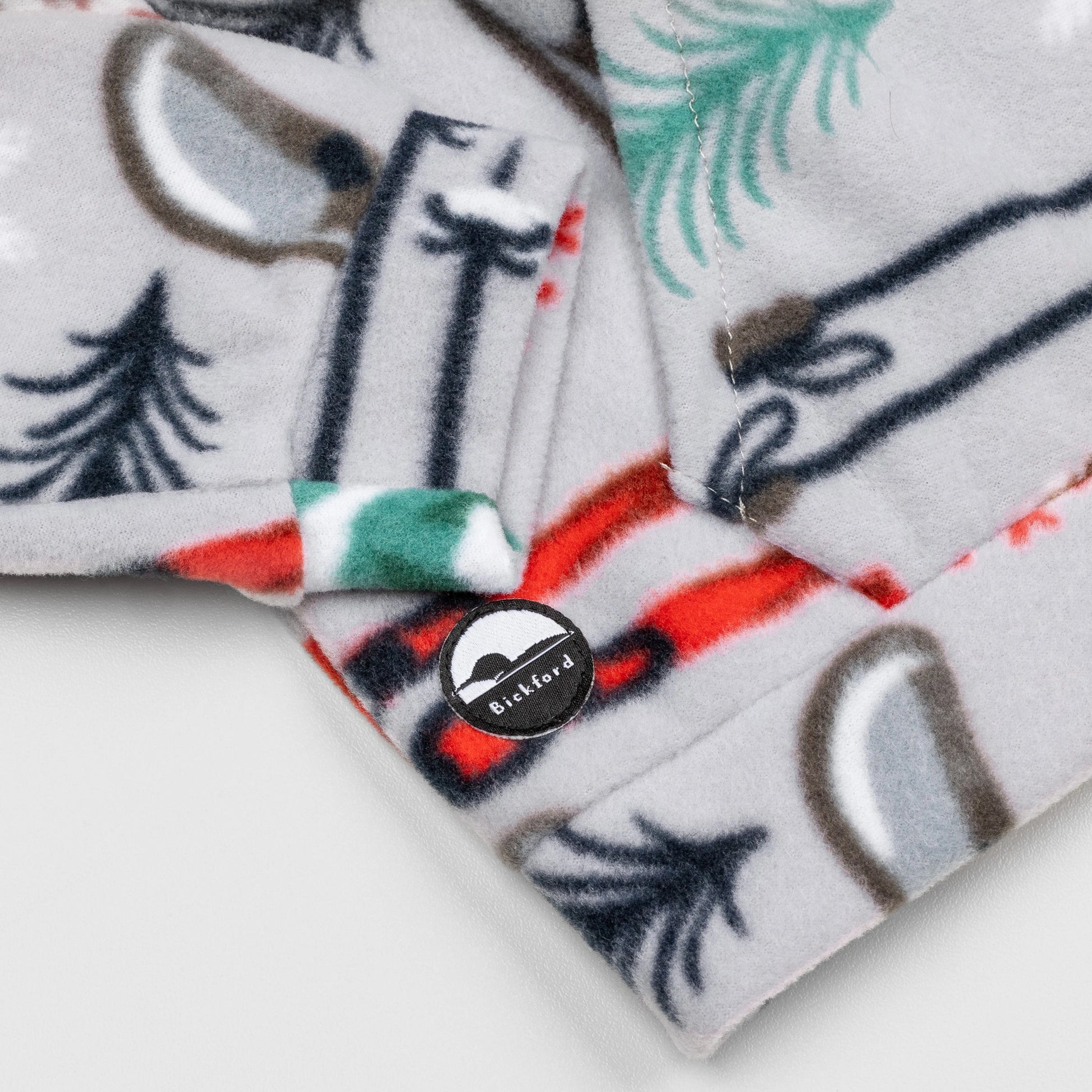 Close-up of the lower hem corner of the Sugarbush Youth Quarter-Zip showing the circular woven Bickford logo patch with a mountain graphic, set against the winter sports-themed print with skis, poles, and pine trees.