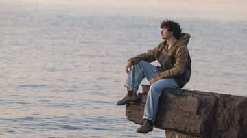 Man wearing Bickford USA the Anorak Sailcloth Jacket, sitting on a rock overlooking Lake Champlain at sunset.