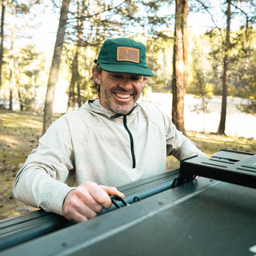 Man wearing a Bickford USA Field and Patch 5 Panel Wool Cap and Athletic Hooded Quarter Zip Pullover, smiling outdoors in a wooded setting.
