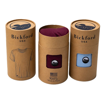 Three Bickford USA kraft cardboard tube tee packaging cylinders arranged in a row against a white background — the left and right tubes are sealed with "Bickford USA" branding and an illustrated t-shirt graphic, the center tube is open showing a burgundy tee inside with a window cutout and "100% Made in USA" American flag stamp, and the right tube has a window showing a light blue tee with a Bickford logo patch.